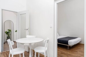 Ein Zimmer mit einem runden weißen Tisch, zwei weißen Stühlen, einem Bett mit dunkler Decke und einem Blumentopf.