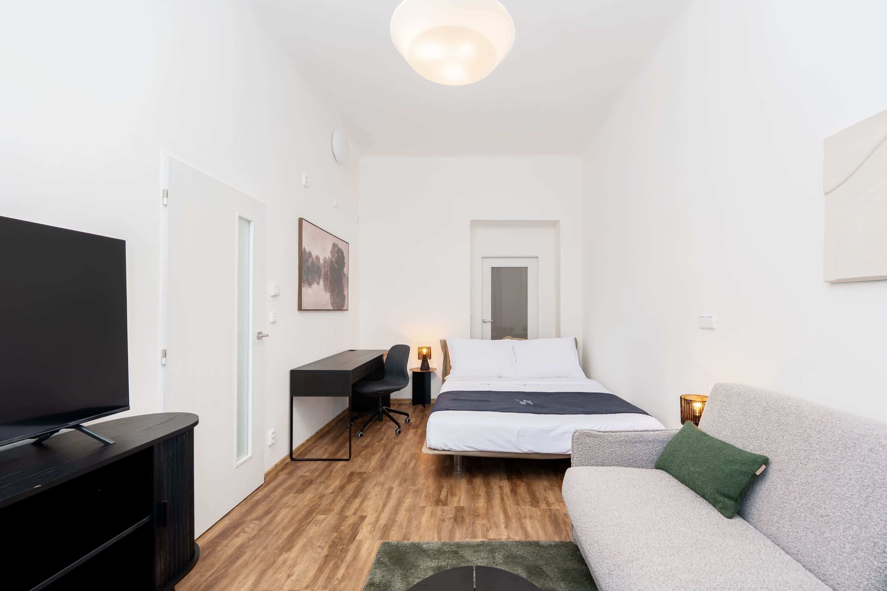 A modern, compact room with a flat-screen TV, a desk with a chair, a bed with a mattress and pillows, and a sofa with a cushion.