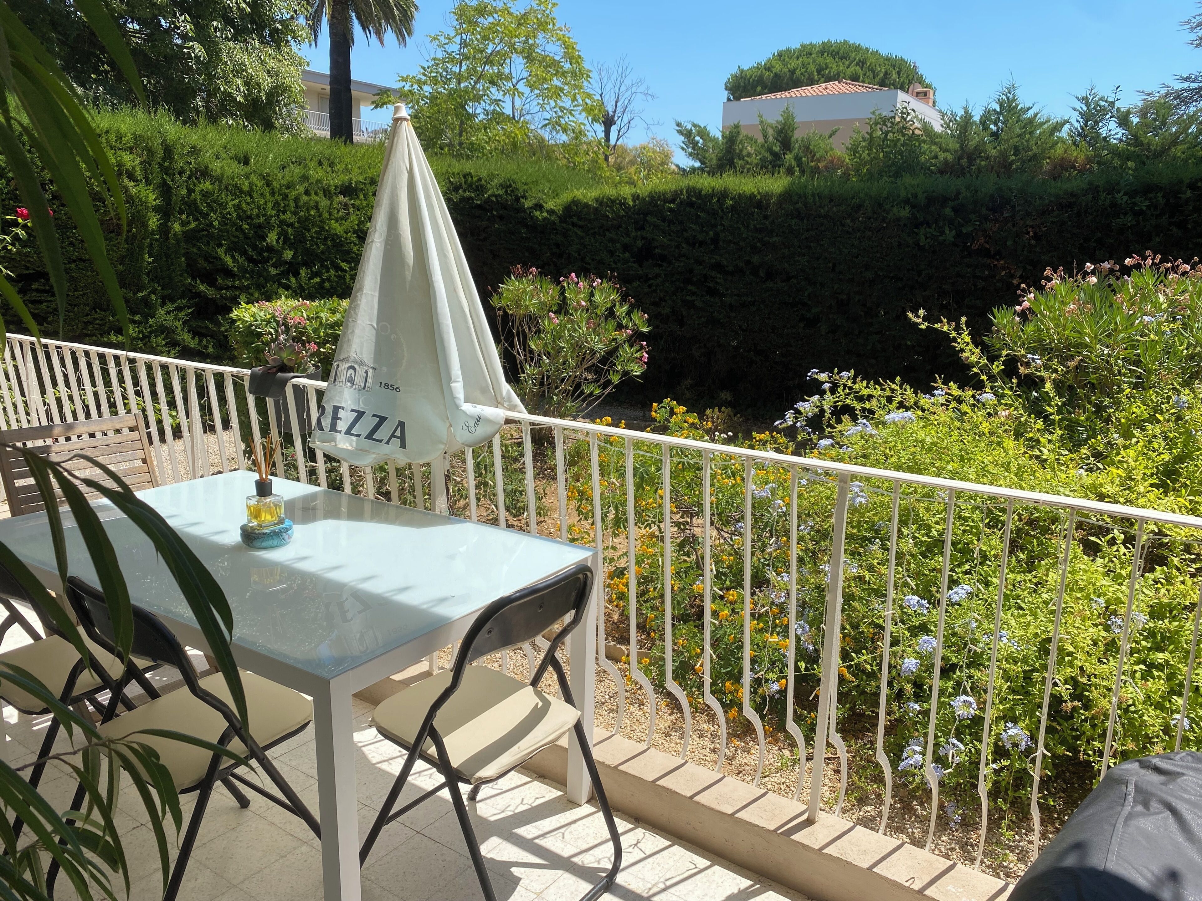 An outdoor seating area with a table and chairs, a white umbrella, and a view of a garden with various plants and flowers.