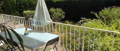 An outdoor seating area with a table and chairs, a white umbrella, and a view of a garden with various plants and flowers.
