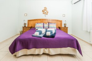 Un lit avec une tête de lit en bois, une couvre-lit violette et plusieurs oreillers. Il y a deux tables de chevet avec des lampes et un luminaire fixé au mur.