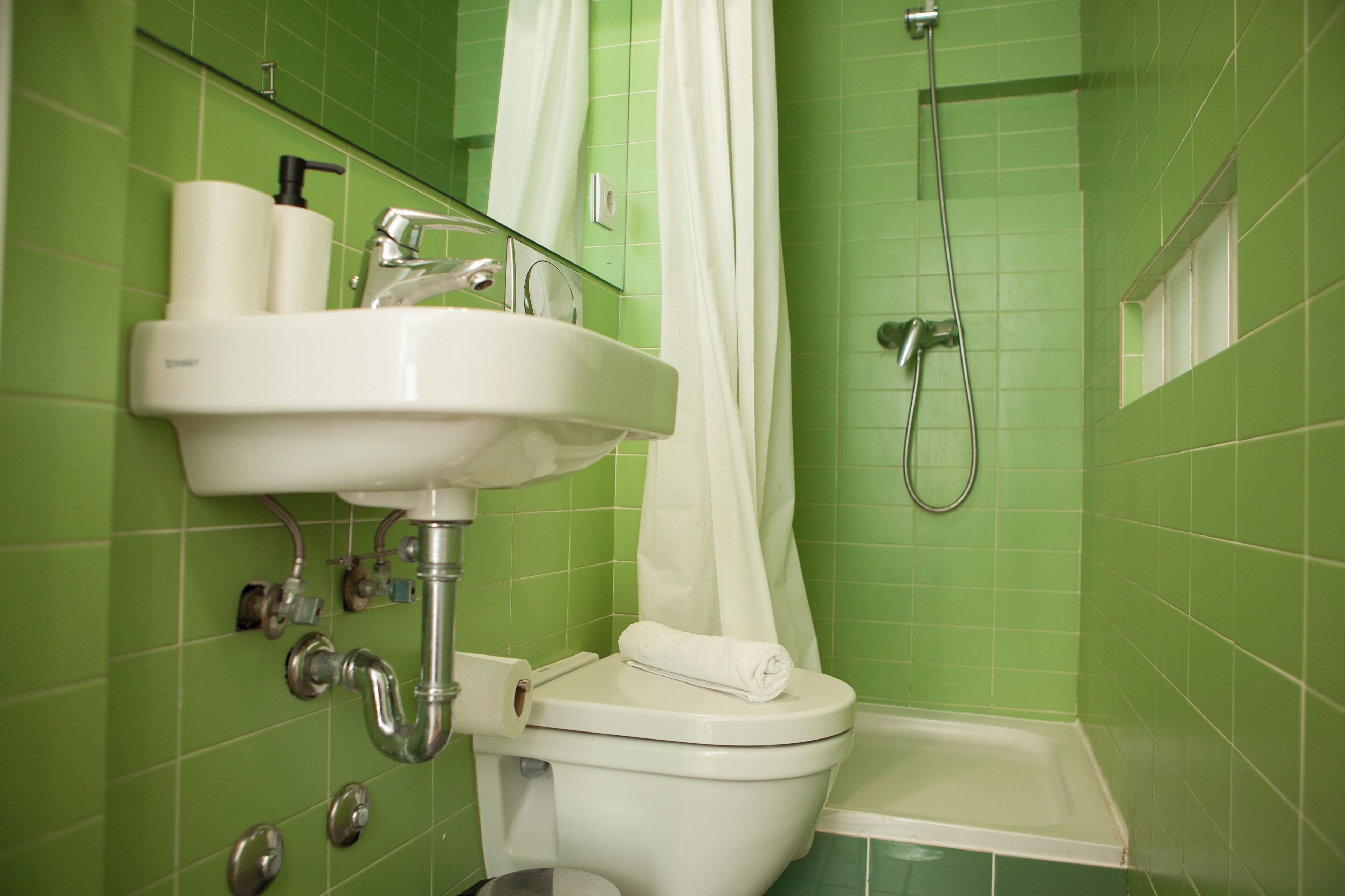 Un bagno con una parete piastrellata in verde, un lavandino bianco con specchio, un water e una zona doccia.
