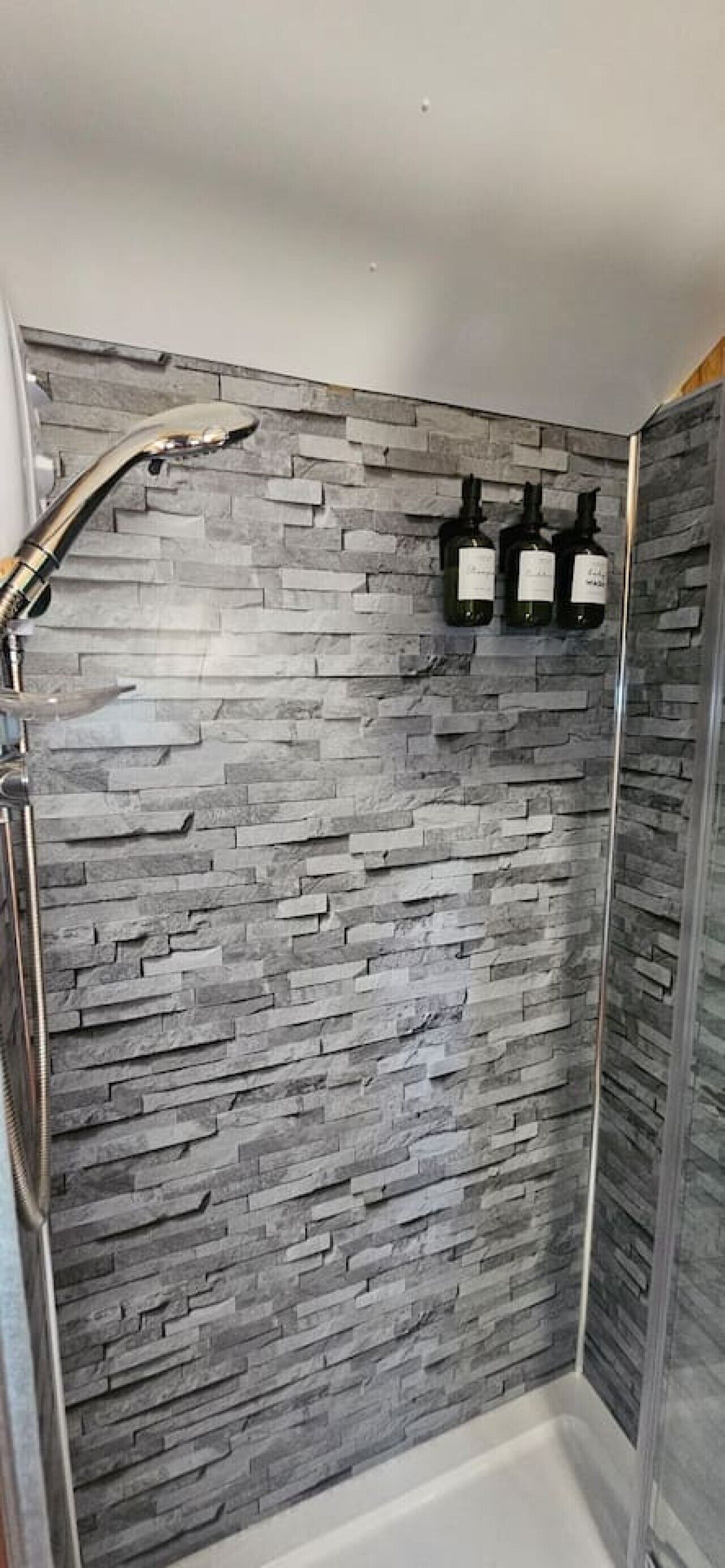 A shower with a stone wall, a glass door, and three bottles on the wall.