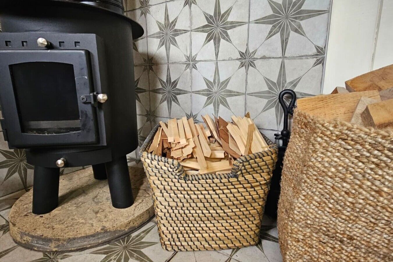 A wood-burning stove, a woven basket with firewood, and a woven basket with a hook.