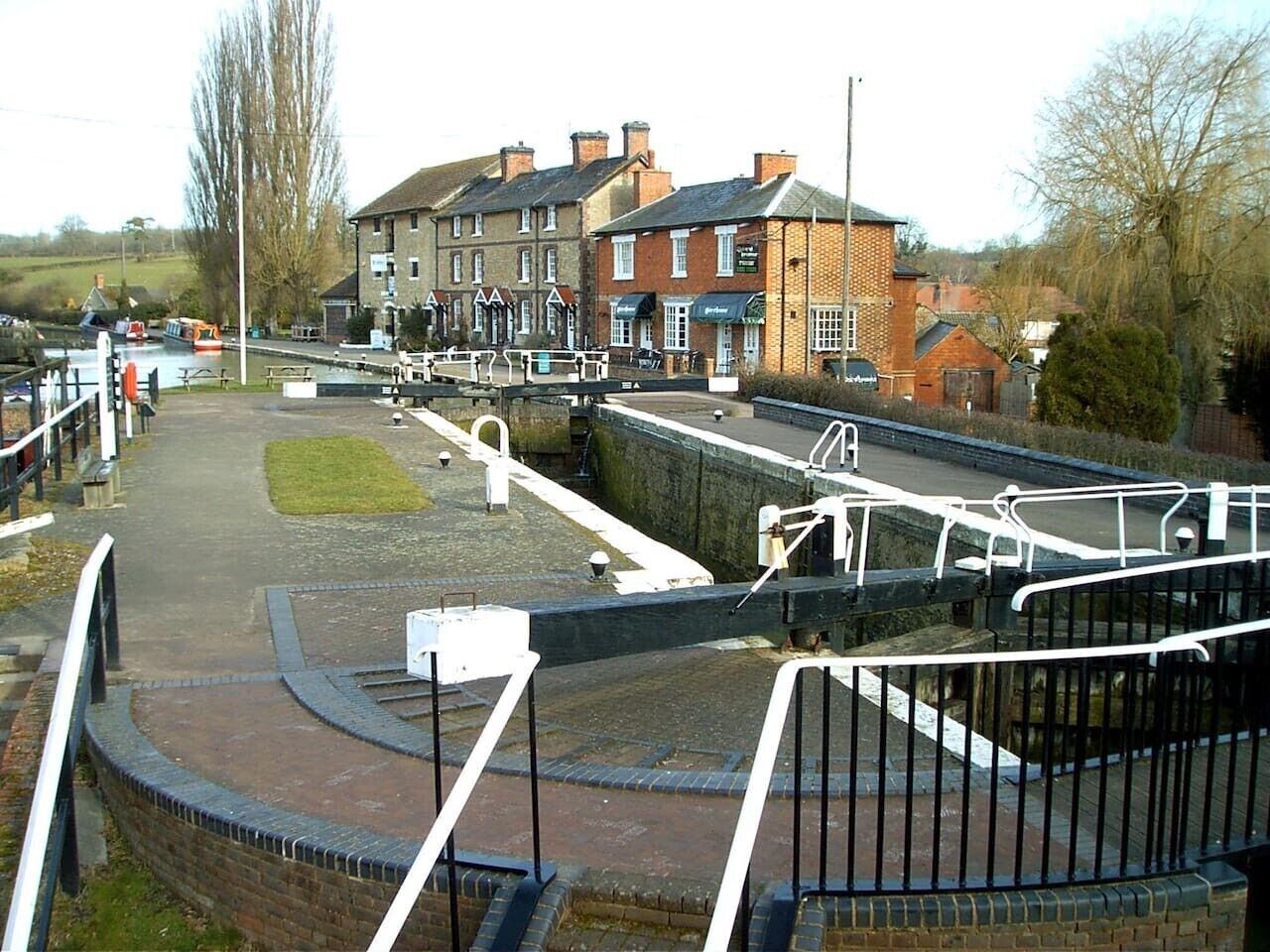 Een pad langs de rivier met een gebouw van baksteen, een brug en een sluis.