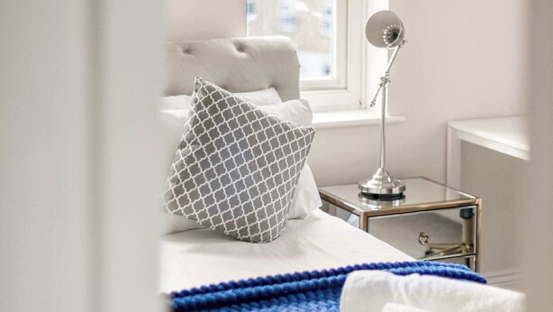 A bedroom with a bed, a bedside table, a lamp, and a window. - Framlingham Court, Romford, London (Greater London)