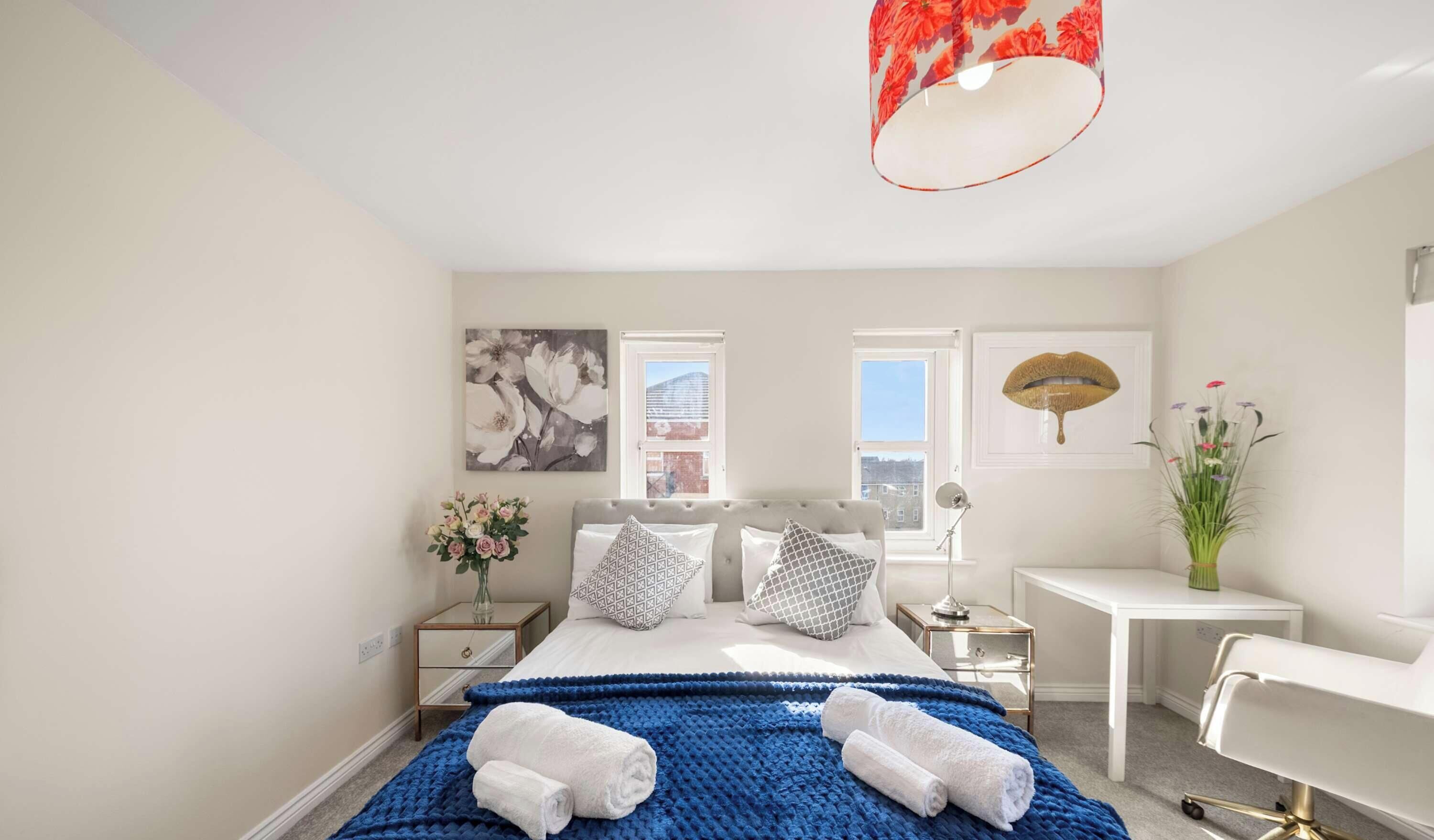 A bright bedroom features a white king bed with a blue blanket, windows, a bedside drawer, and a small workspace.