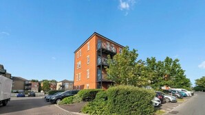 A multi-story residential building with a brick facade, a balcony on the upper floor, and a parking area with cars. - Framlingham Court, Romford, London (Greater London)