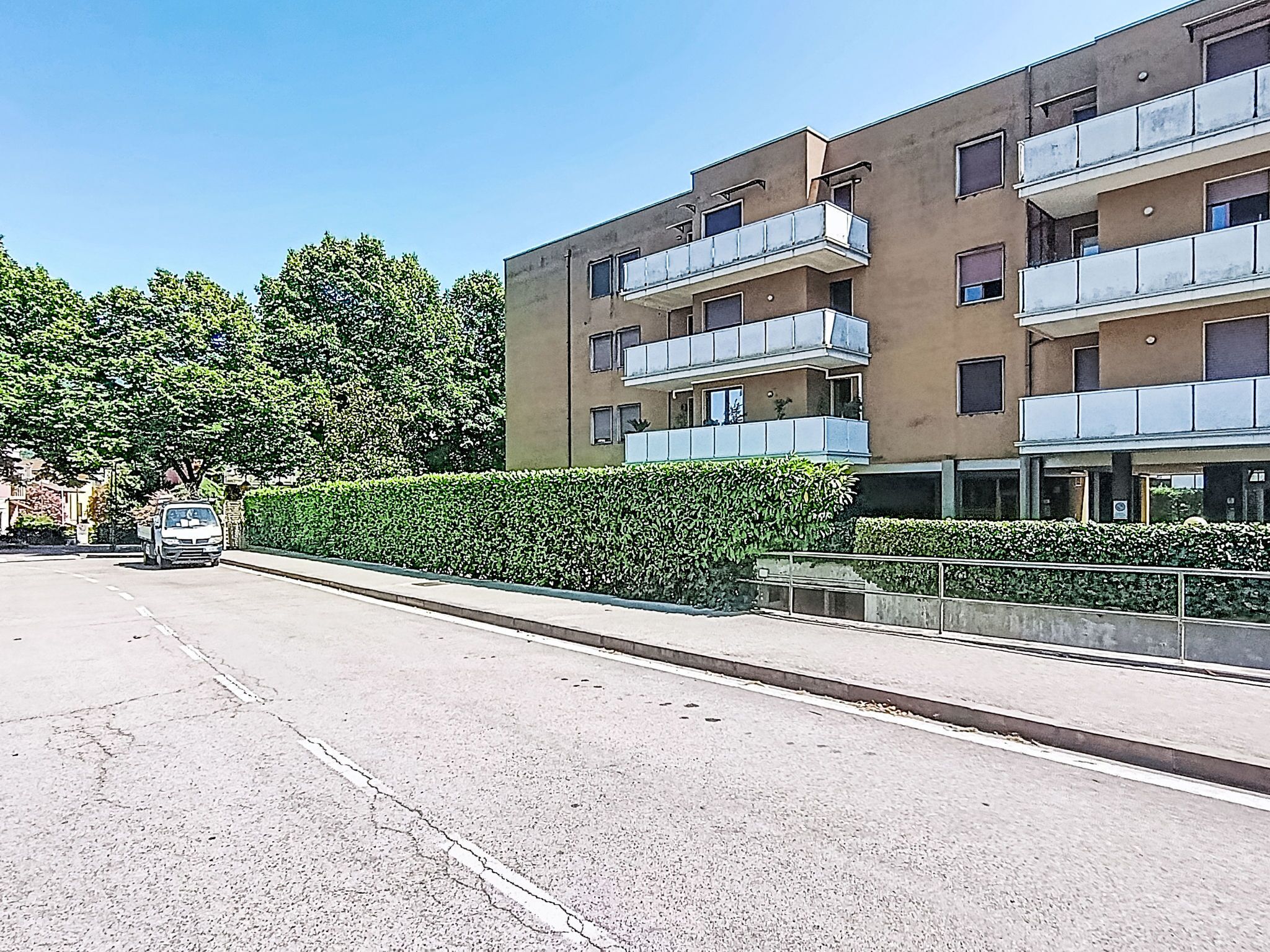 A multi-story residential building with balconies, a paved road, and a hedge.