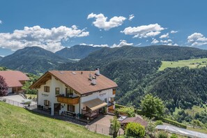 A two-story chalet with a balcony, a patio with outdoor seating, and a view of the surrounding mountains. - Family-Friendly Apartment “Alpe Unterkehrhof” with Mountain View, Garden & Terrace (Lajen)