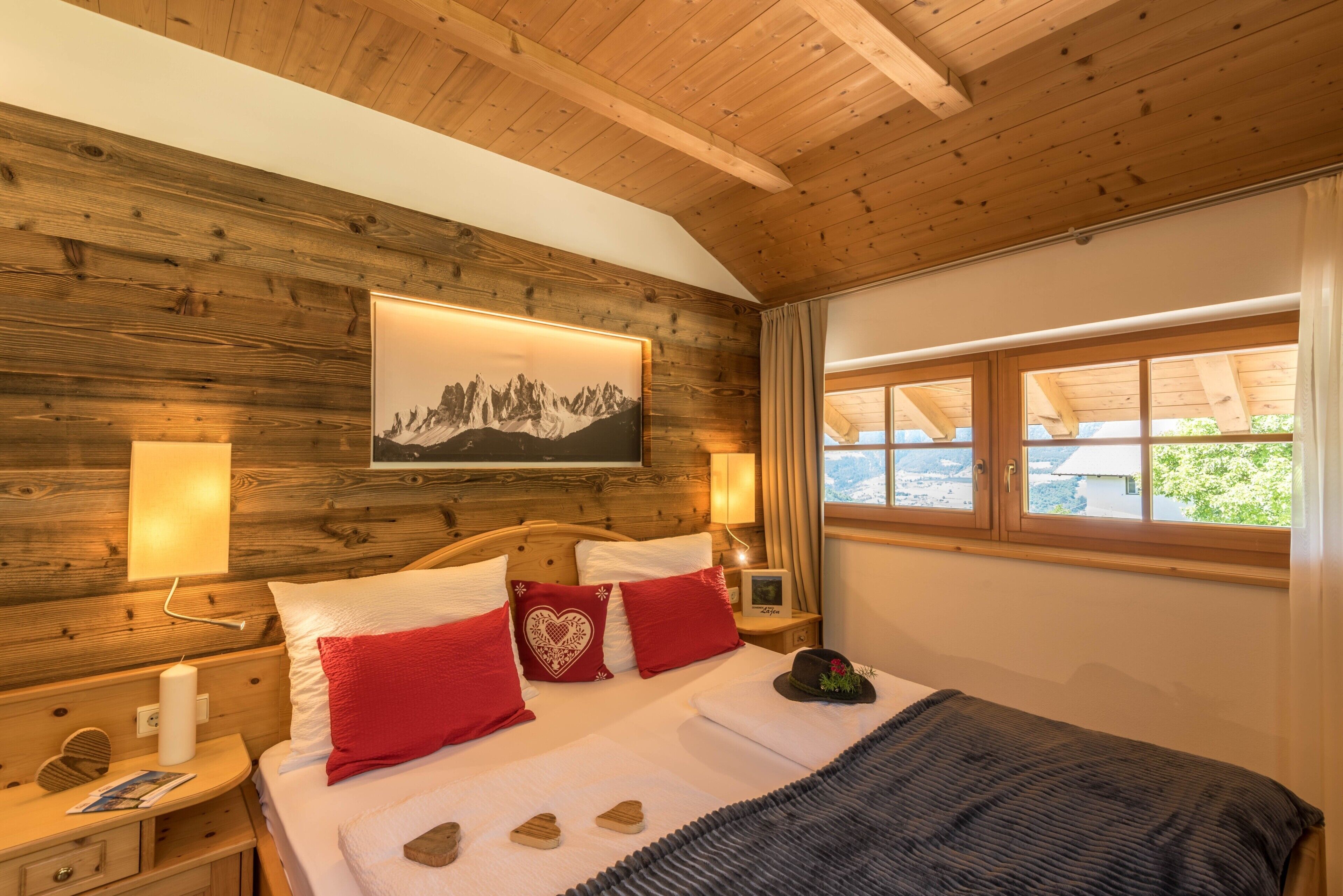 Een slaapkamer met houten lambrisering, een bed, rode kussens, een berglandschap op een schilderij en een raam met uitzicht.