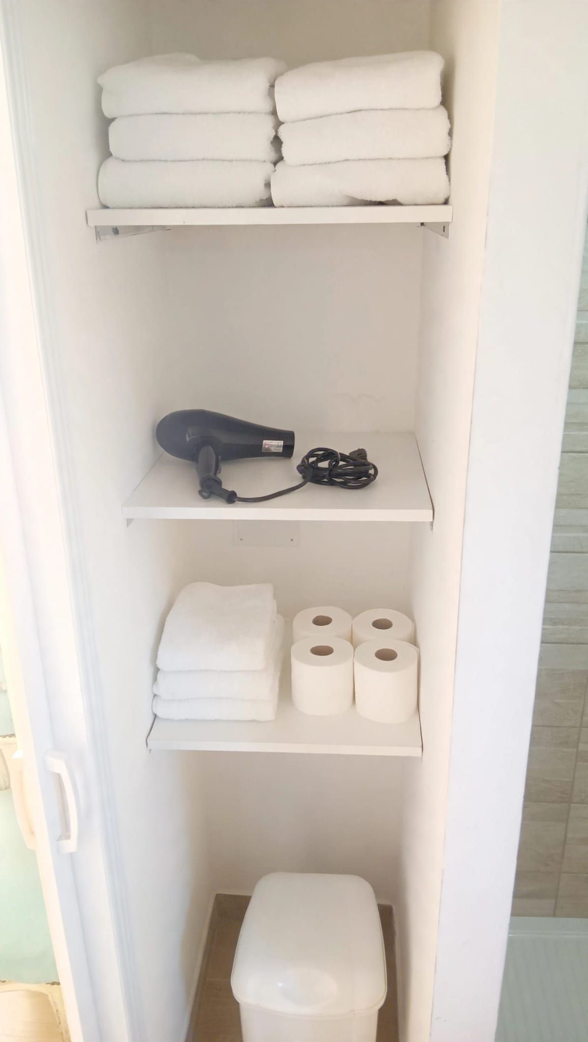 A bathroom with a toilet, a shelf with towels, a hairdryer, and toilet paper.