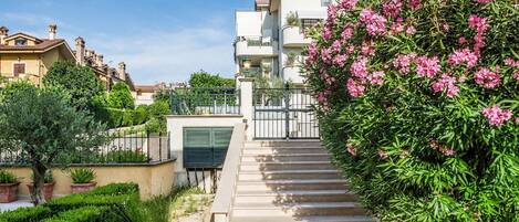 A residential building with a staircase leading to a gated entrance, surrounded by lush greenery and flowering plants.