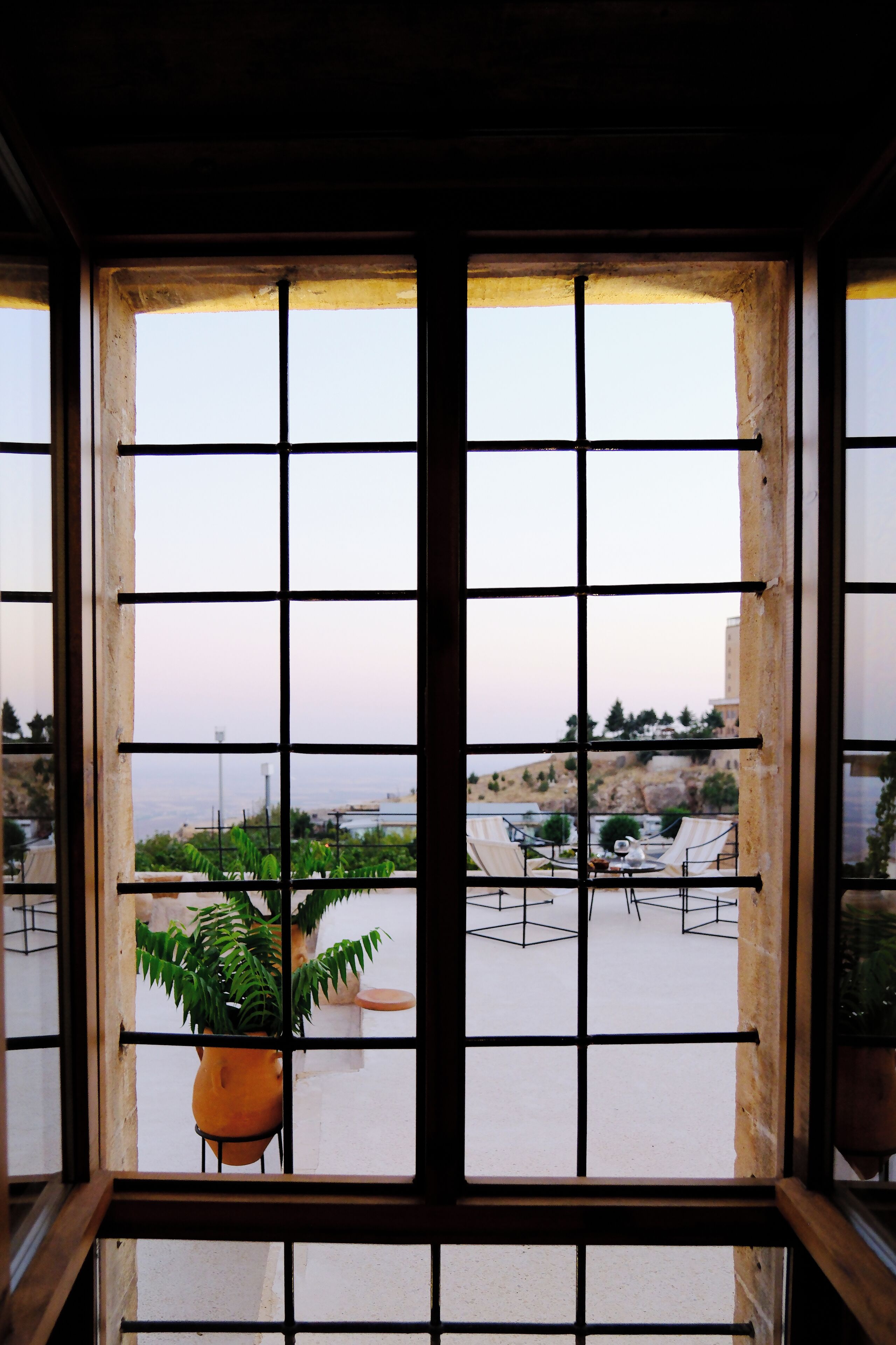 A window with a grid pattern, through which a patio area with chairs and a potted plant is visible.