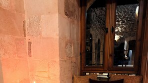 Un muro de piedra con una vela sobre un soporte, un marco de ventana de madera y un banco acolchado con un cojín oscuro.