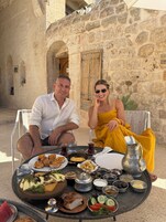 A couple seated at a table with a variety of food items, including bread, salads, and dips, in front of a stone building with a wooden door.