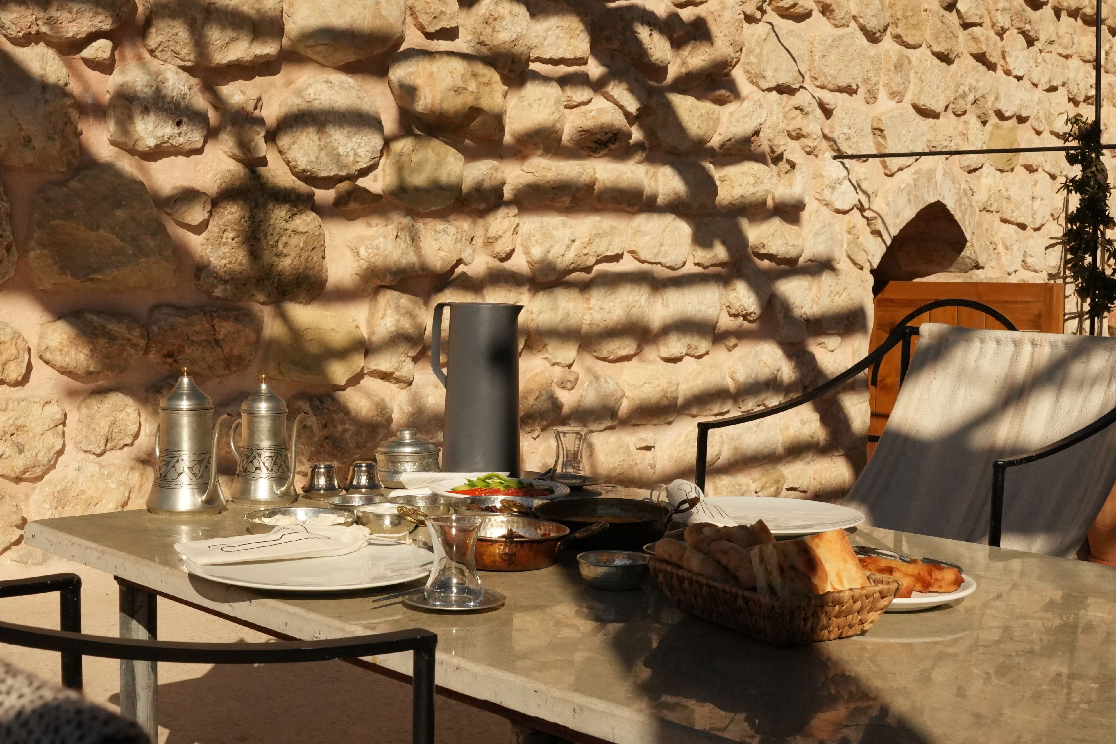 Une table dressée à l’extérieur, avec un mur de pierres en arrière-plan, agrémentée d’un pichet en métal, d’assiettes, de verres et de pain.
