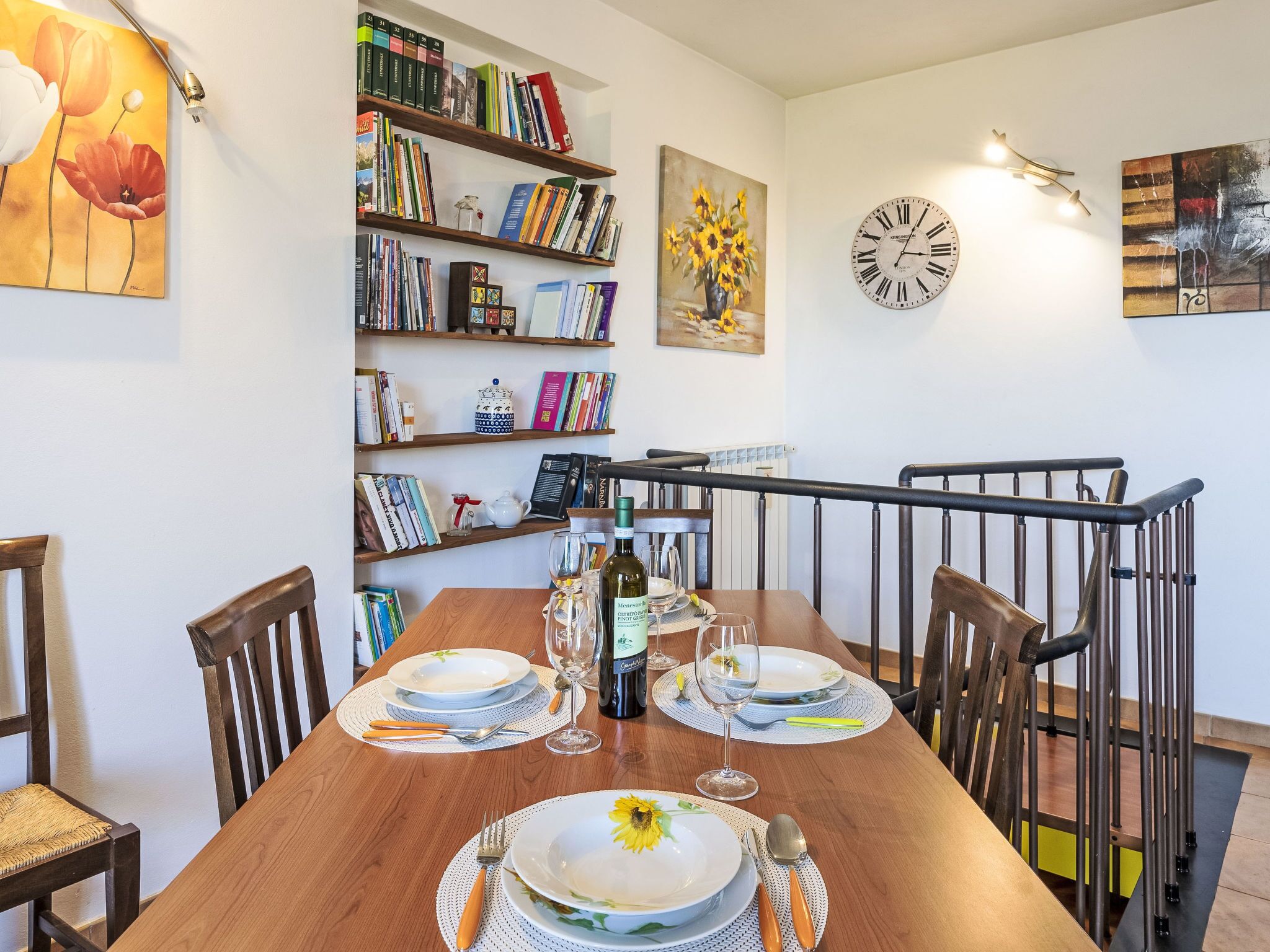A dining area with a wooden table set for two, bookshelves with books, a clock, and artwork on the walls.