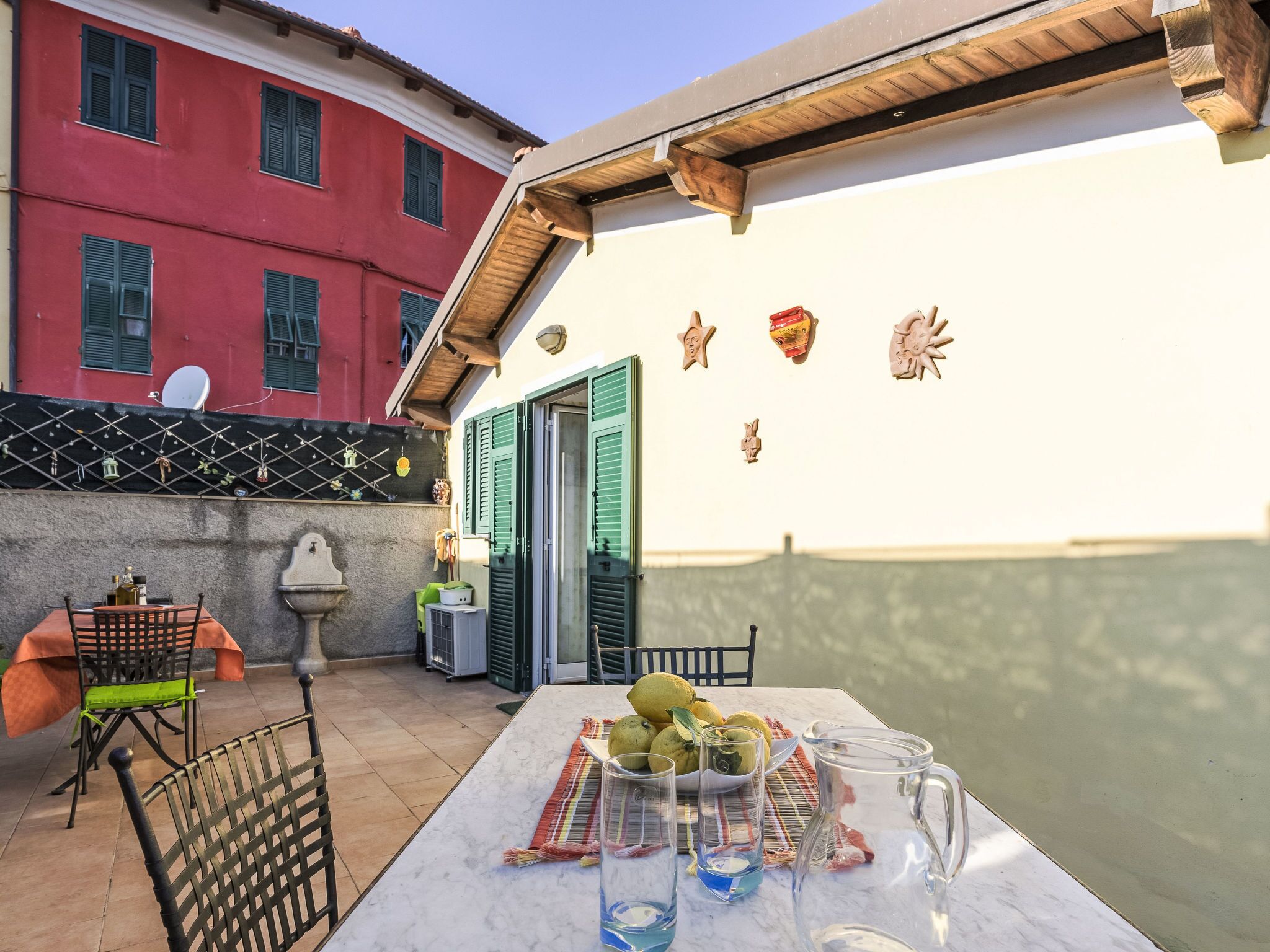 A patio with a table set for dining, a chair, a small table with a bowl of fruit, and a pitcher.