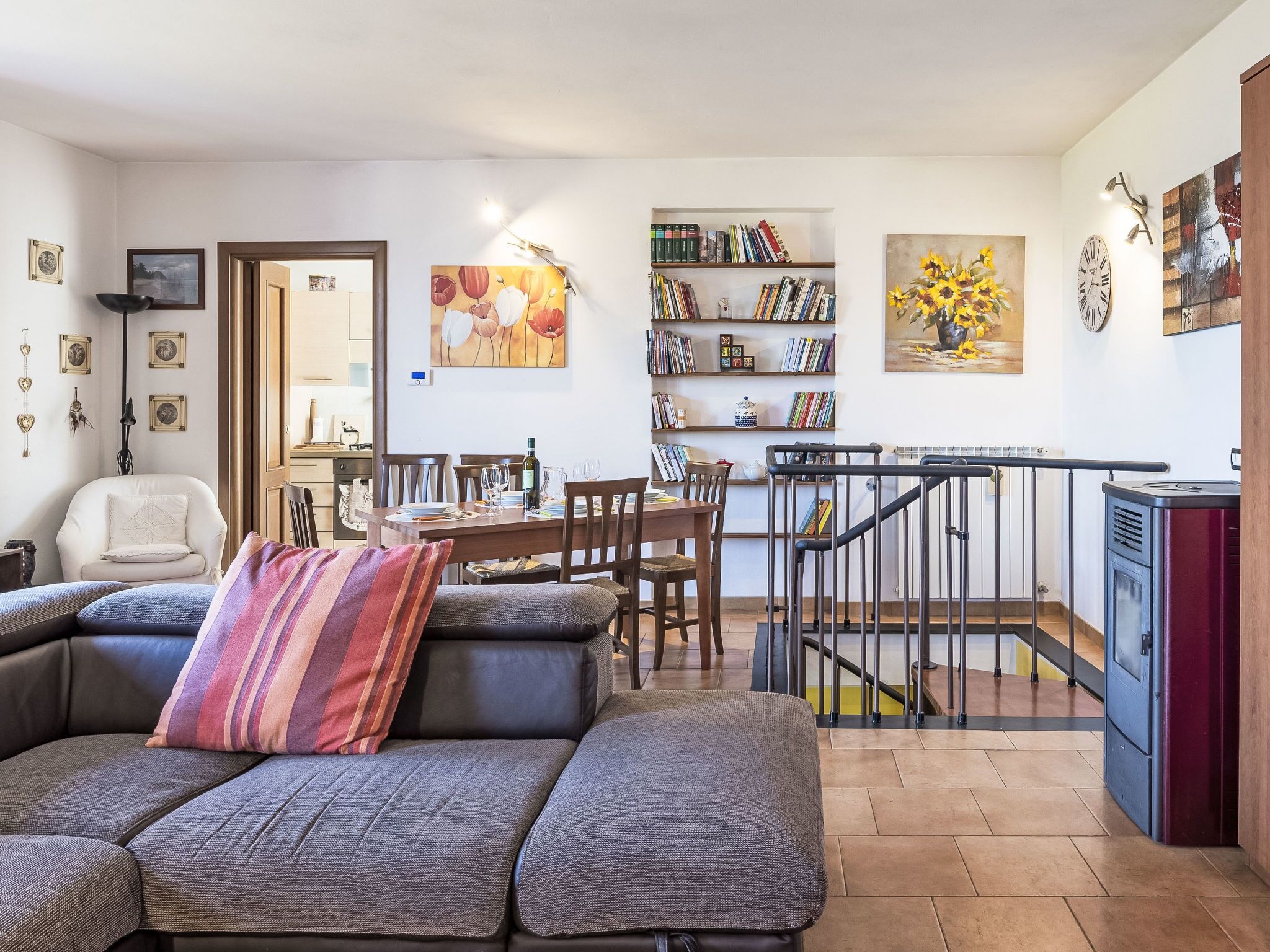 A modern living room with a sectional sofa, a dining area with chairs, a bookshelf, and a staircase with a railing.