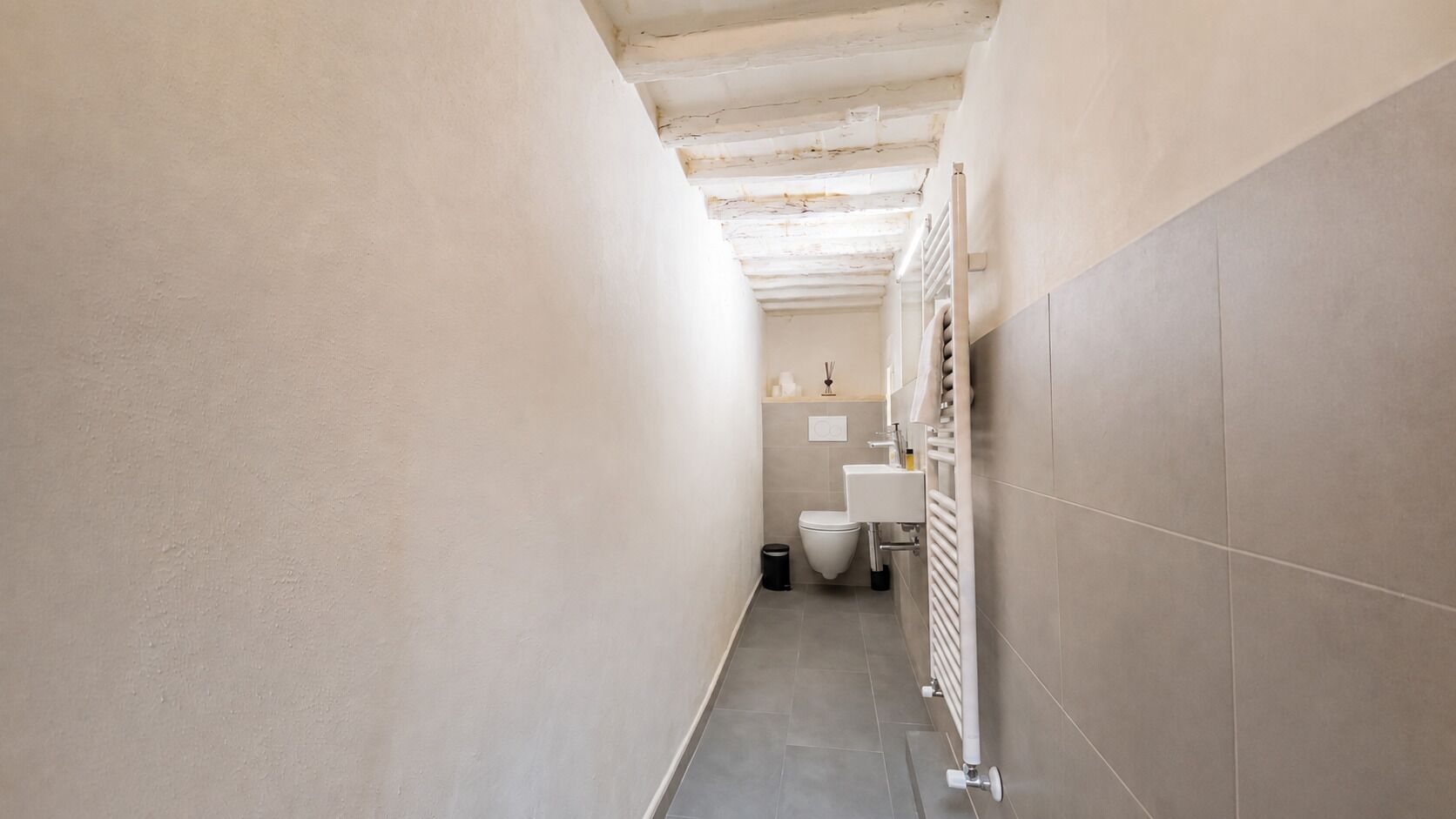 Un pasillo estrecho con un inodoro y un lavabo a la derecha, un radiador a la izquierda y suelo de baldosas.