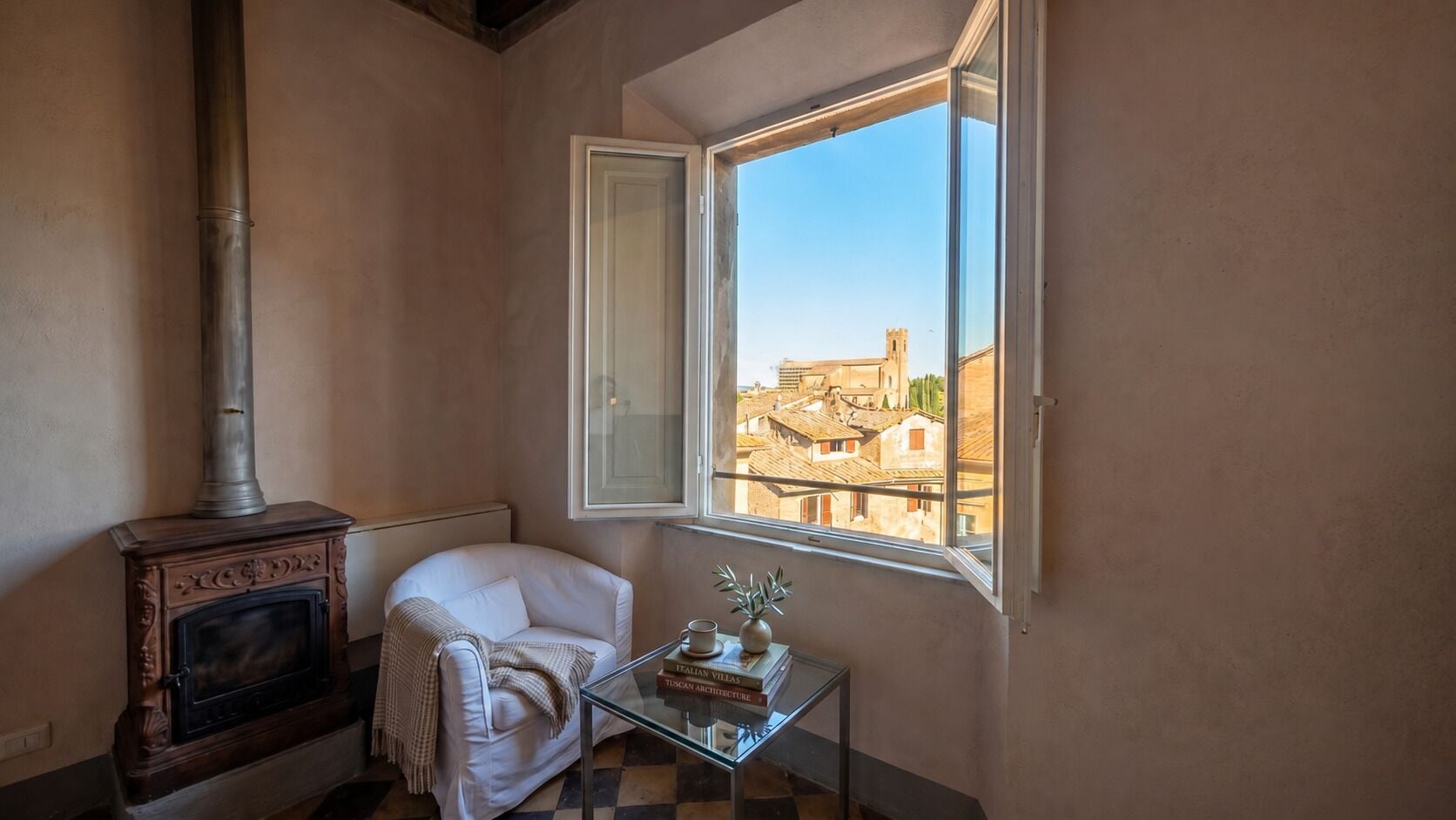 Une pièce avec une cheminée, un fauteuil blanc, une petite table avec des livres et un vase, et une fenêtre donnant sur des immeubles.