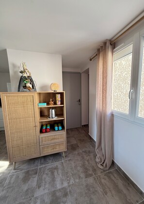A room with a wicker cabinet, a window with curtains, and a door leading to another room. - Les Roses by Alto Service (Le Barcarès)