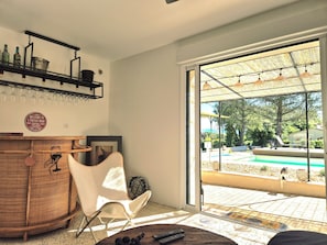 A room with a bar area, a chair, a table, and a view of an outdoor swimming pool. - Le Repos House—Sleeps 6—Pool—Pétanque (Alleins)