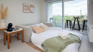 A bedroom with a bed, a small table with a kettle, a chair, and a balcony with a table and chairs. - Corporate Stays First Living - 306 (Asunción)