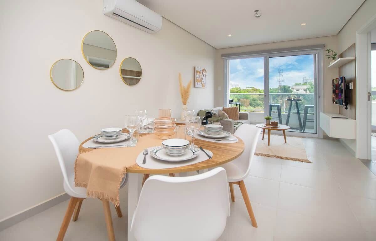 A dining area with a round table, white chairs, and a beige tablecloth. There are three round mirrors on the wall, and a large window with a view of greenery and buildings.