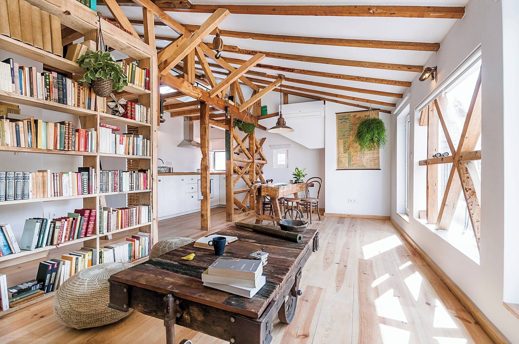 A room with exposed wooden beams, a bookshelf, a wooden table with books, and a large window.