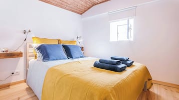 A bedroom with a bed, a bedside table, a lamp, a window, and a wall socket.