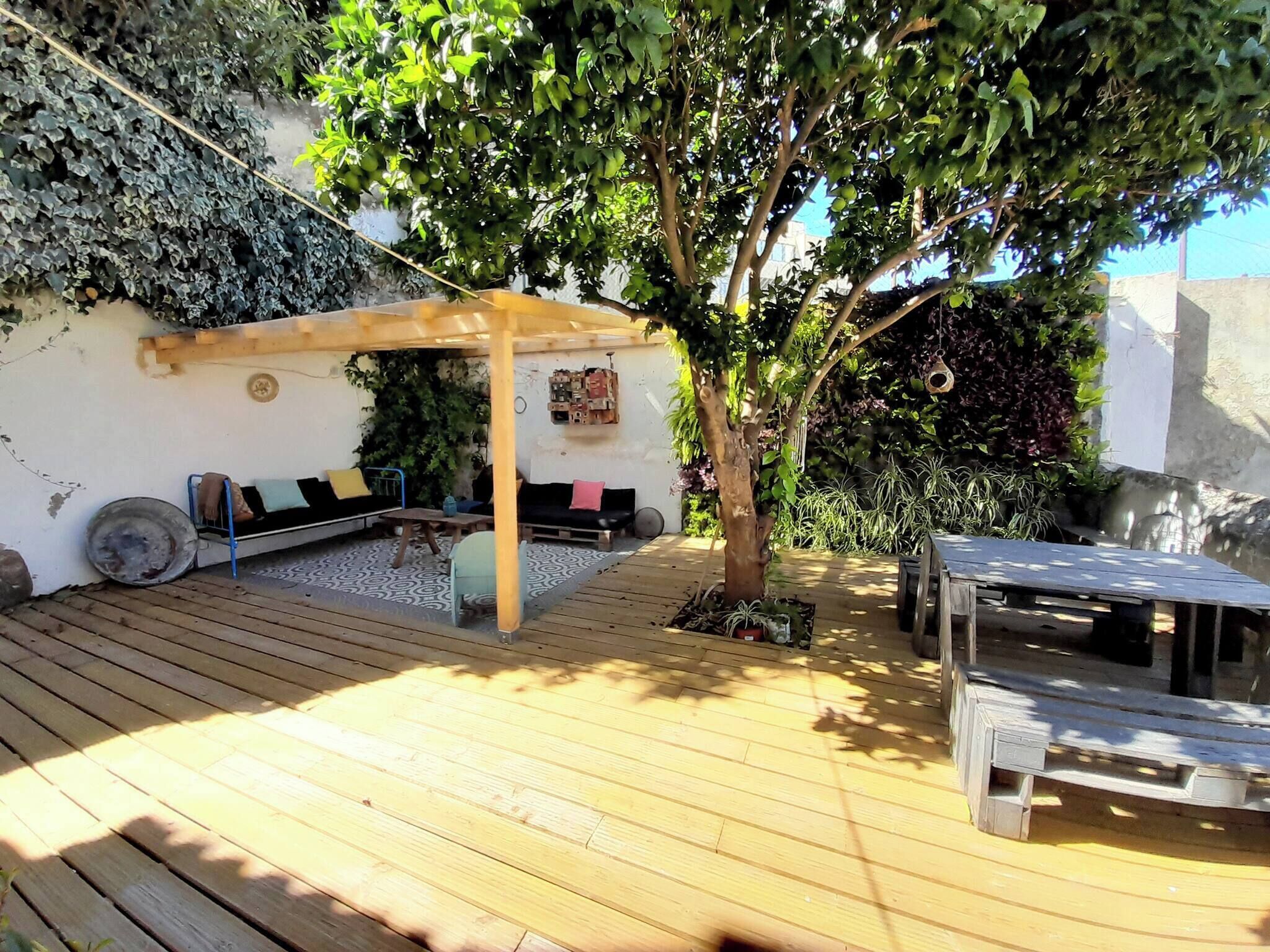 Una terraza de madera con zona de estar cubierta, un árbol y una pequeña mesa.