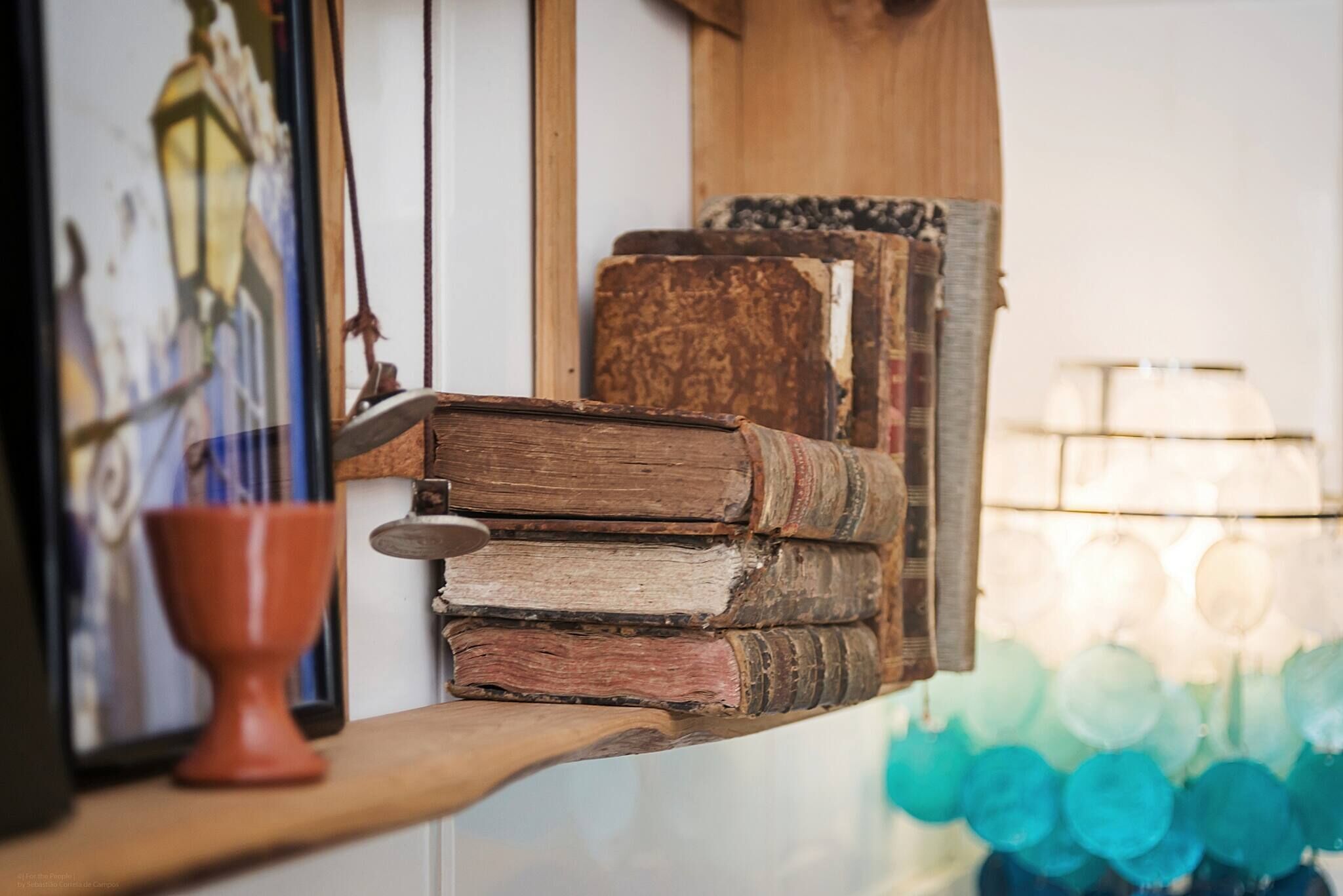 A stack of old books on a wooden shelf, a ceramic cup, and a lamp with a blue glass shade.