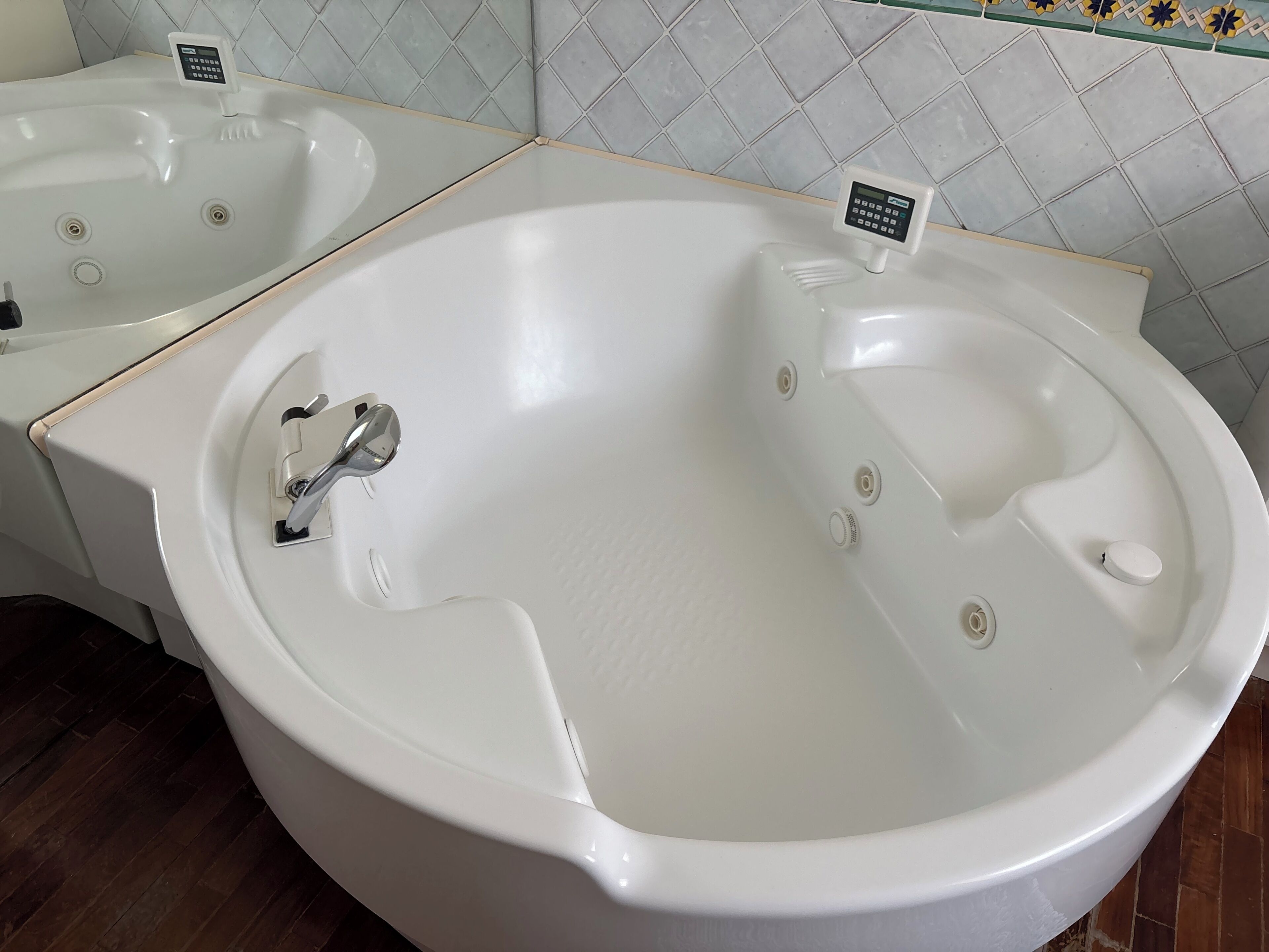 A large, white, freestanding bathtub with a control panel, a smaller sink, and a tiled wall with a diamond pattern.