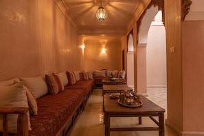 A traditional Moroccan interior with a long sofa, a wooden table with tea set, and a decorative ceiling light.