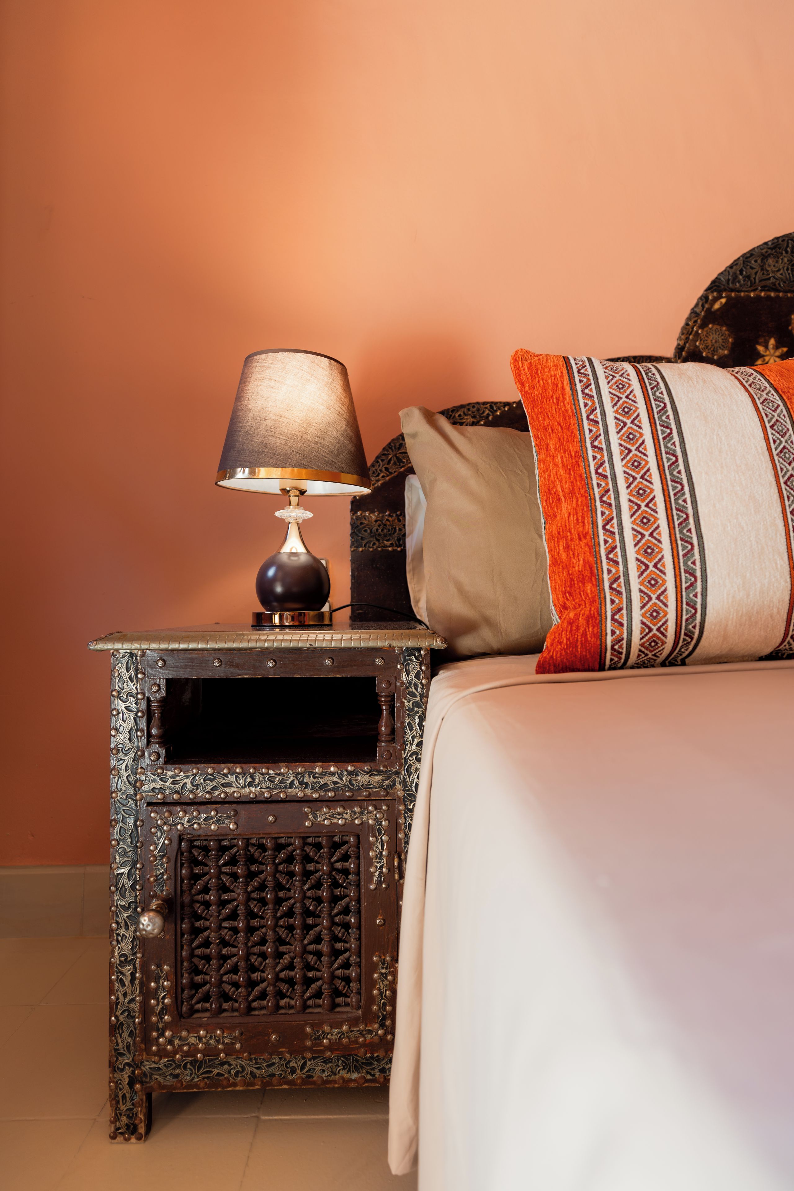 A bed with a patterned pillow, a lamp on a wooden nightstand, and a plain wall.