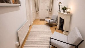 Un couloir avec un radiateur, un canapé gris, une cheminée blanche et un tableau encadré au mur.