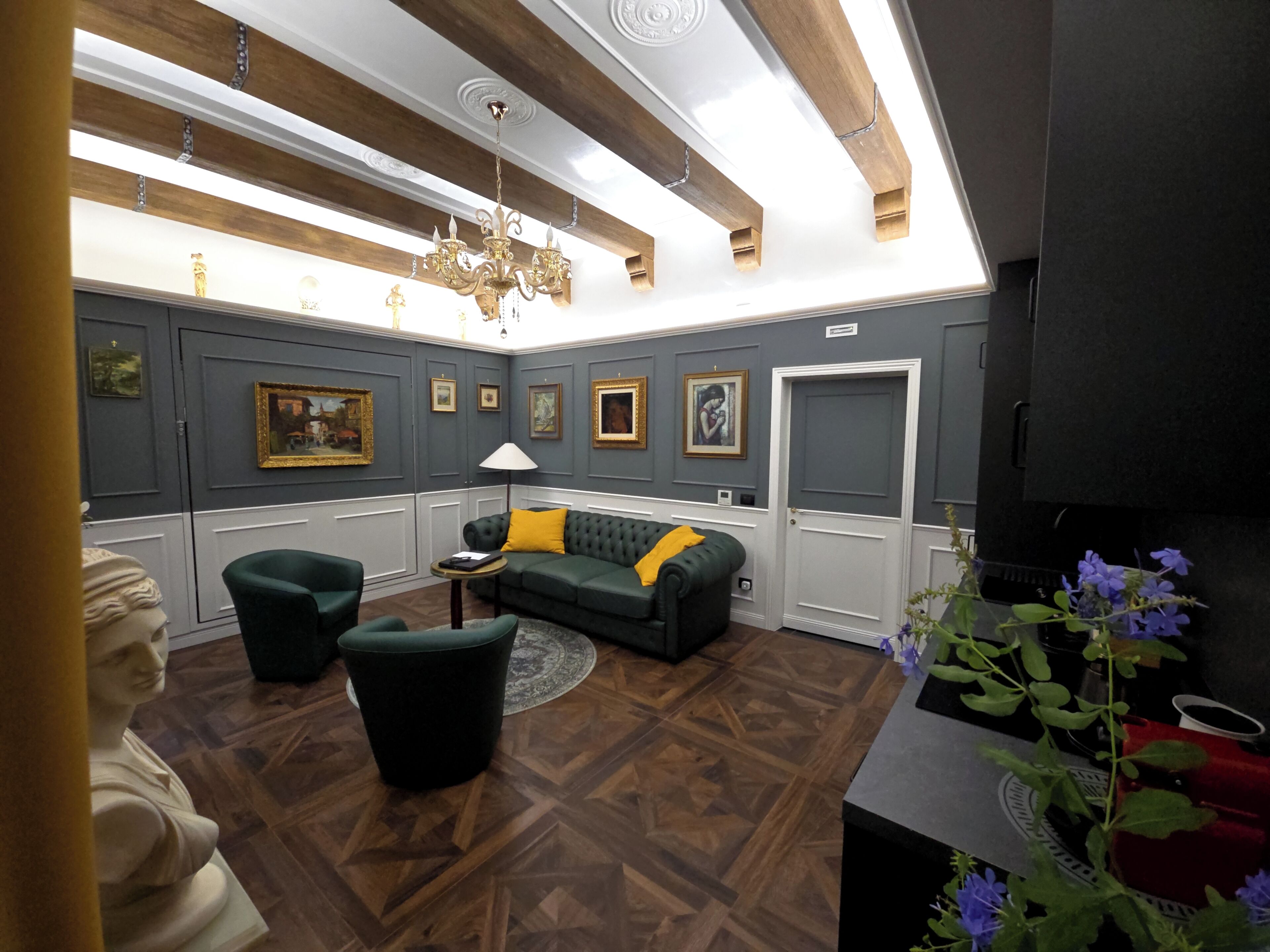 Um quarto com um sofá verde escuro, uma mesinha redonda pequena, um candelabro e uma porta com placa de identificação.