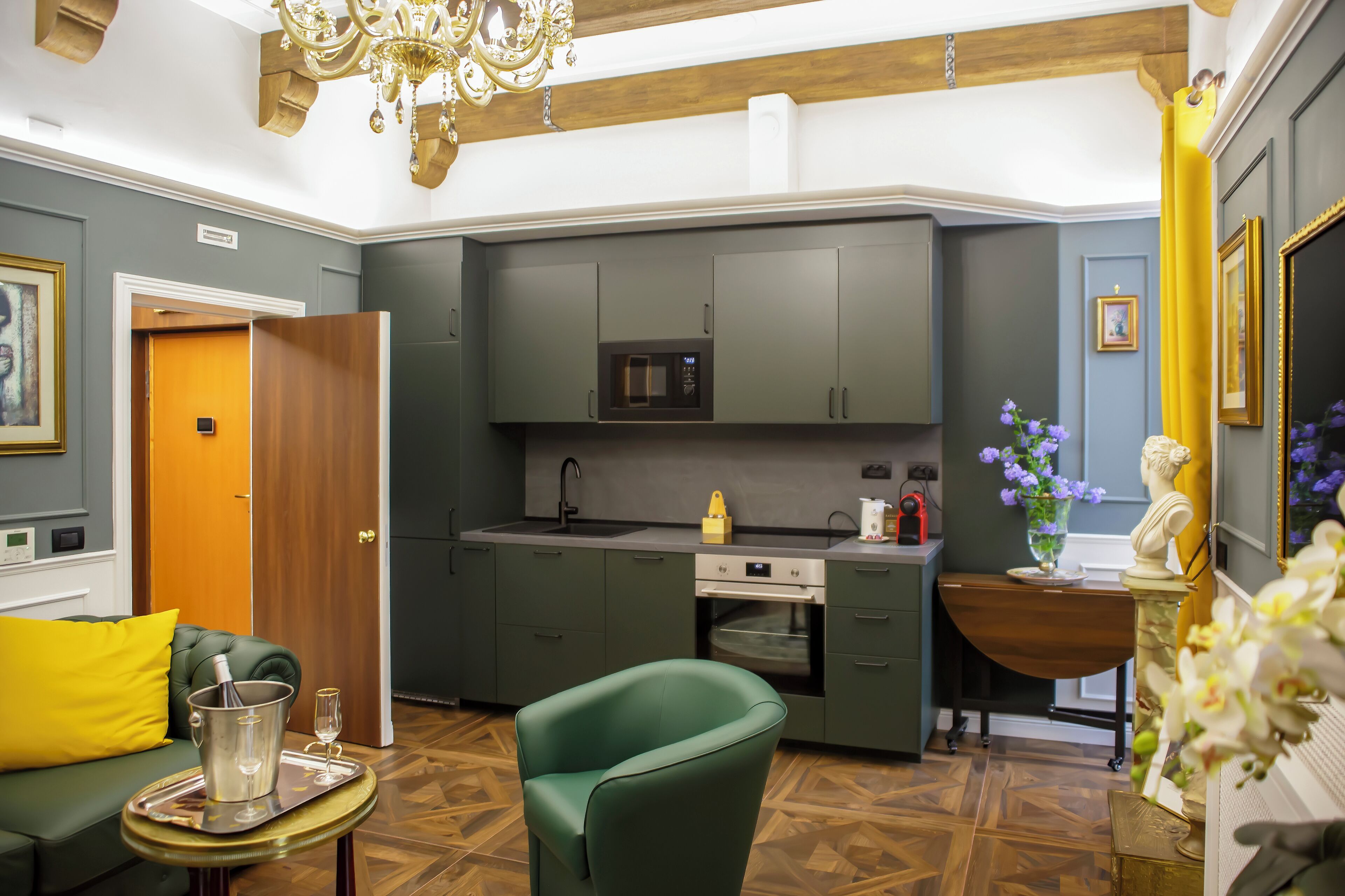 A modern kitchen with grey cabinets, a microwave, and an oven. There is a green armchair, a small table with a tray, and a chandelier.