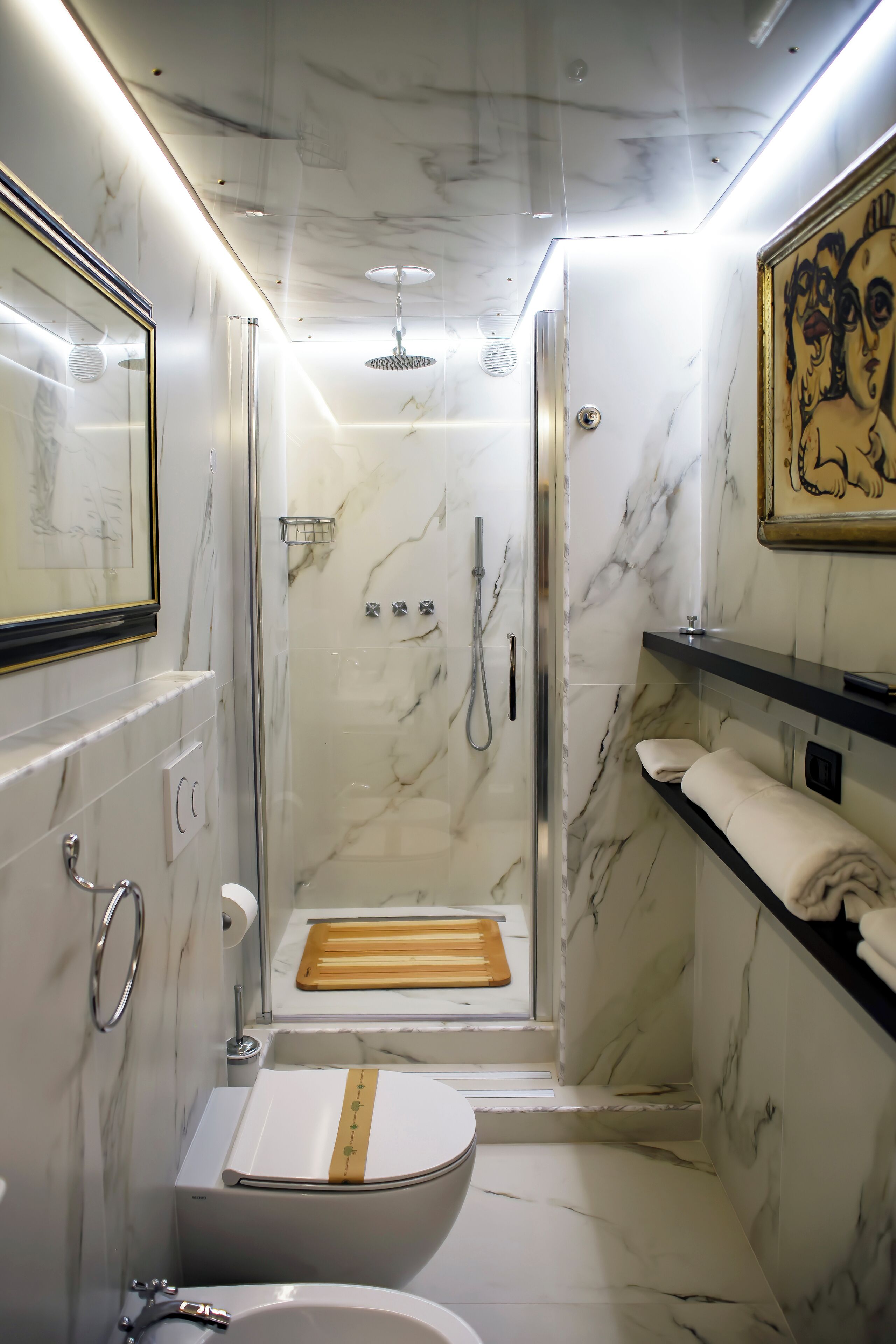 Baño compacto con ducha, inodoro y lavamanos, que presenta paredes de mármol y una obra de arte enmarcada.