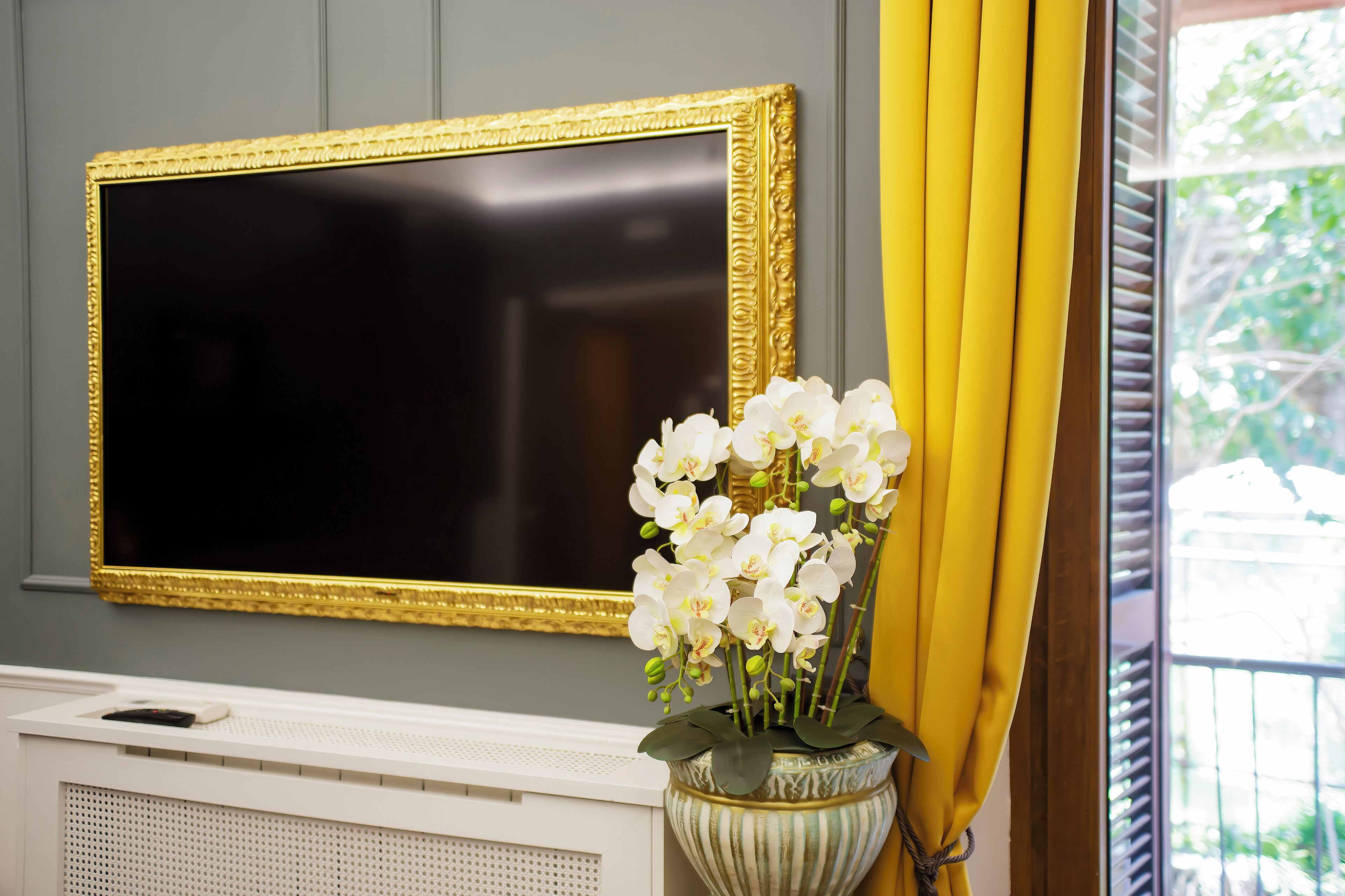 A room with a large framed television, a radiator, and a vase with white flowers.