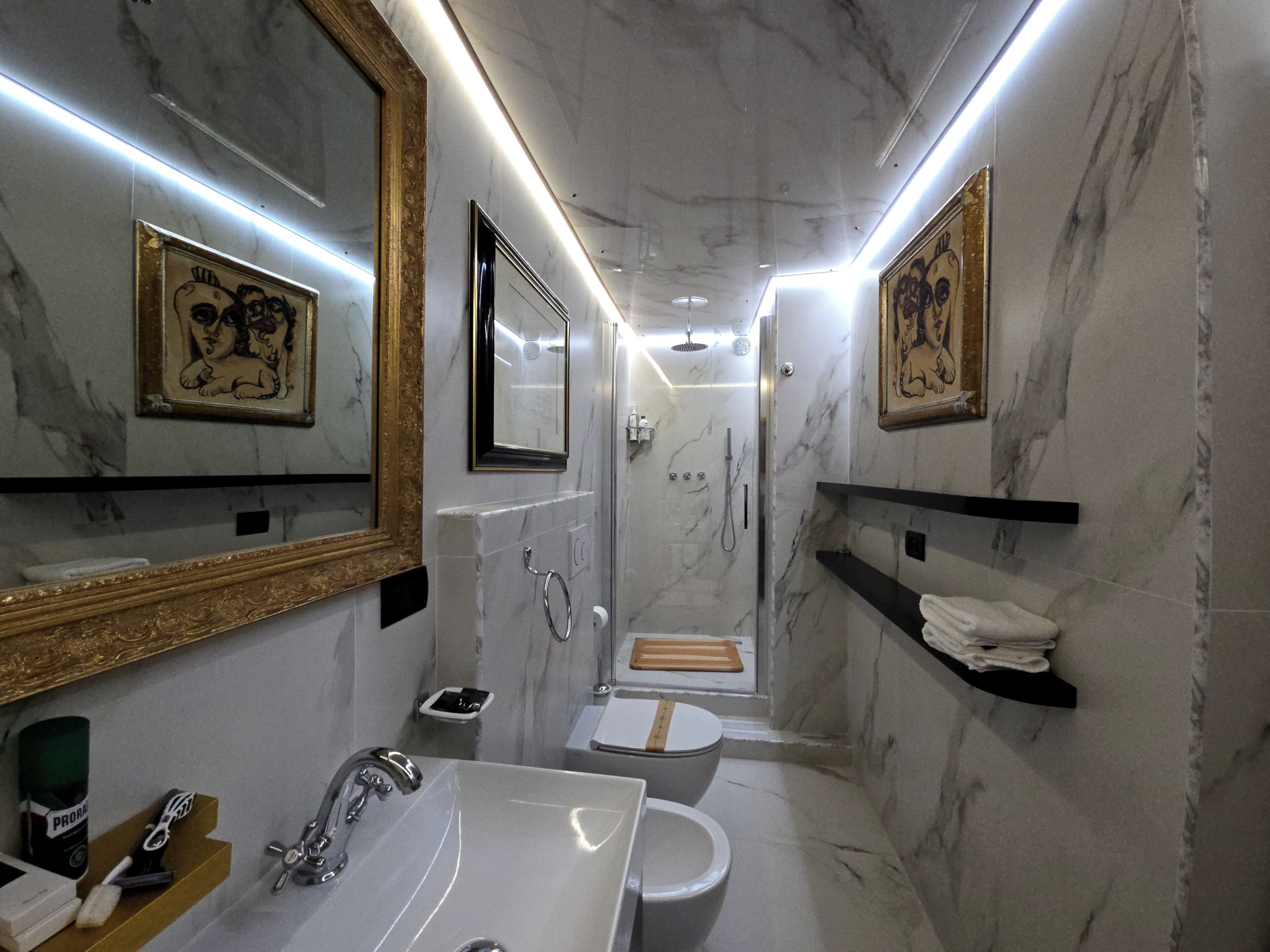 Ein modernes Badezimmer mit Marmorwänden, einem Waschbecken, einer Toilette und einer Dusche. An den Wänden befinden sich Bilder in Rahmen, und Regale sind mit Handtüchern bestückt.