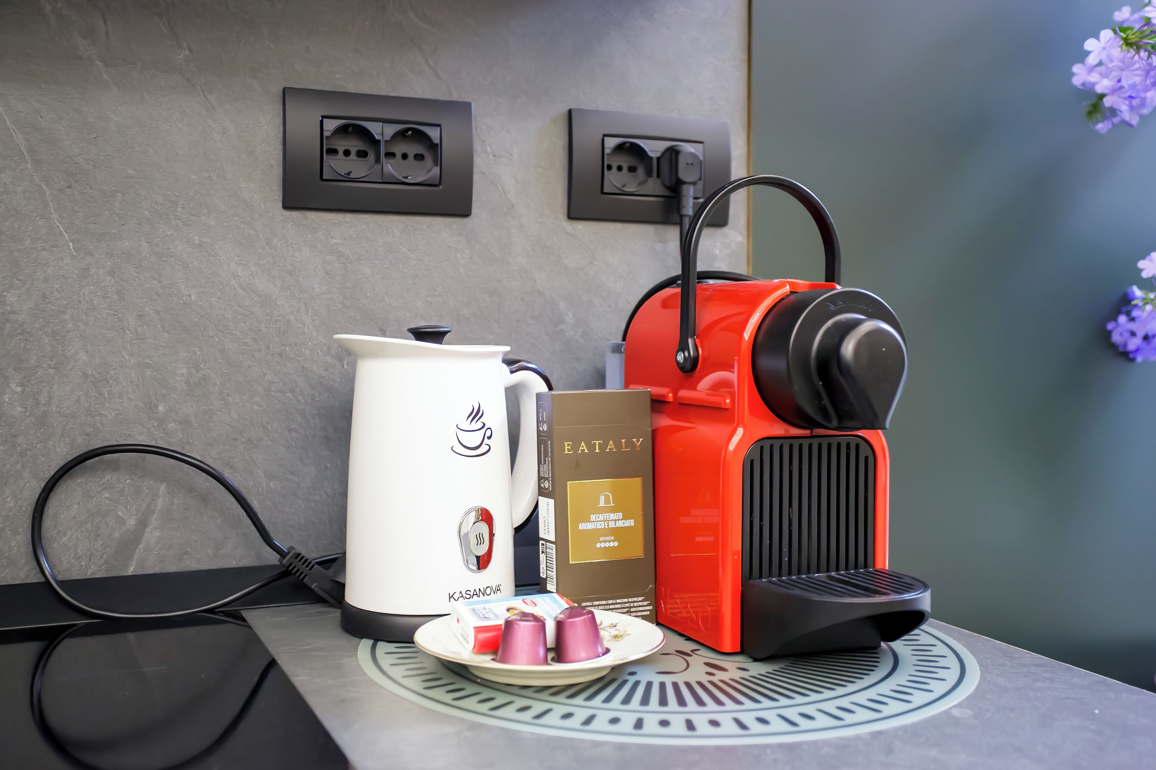 A coffee machine, a white electric kettle, and a plate with cups on a table with a power outlet and a wall socket.