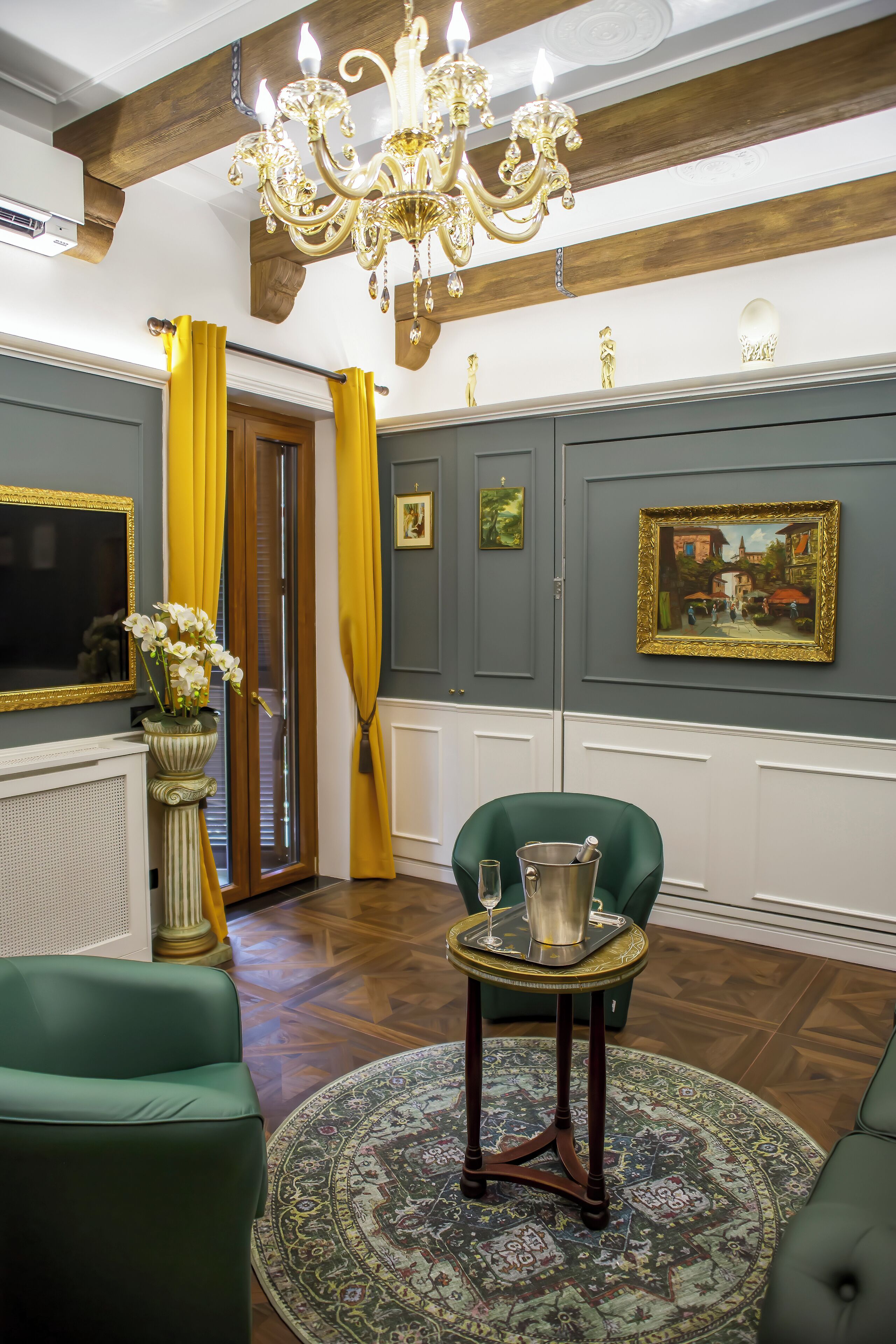Una habitación con una araña, sillones verdes, una mesa redonda con una bandeja y un cuadro grande en la pared.