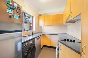 A compact kitchen with wooden cabinets, a built-in dishwasher, a washing machine, and a window with a view of a balcony. - Penthouse with Harbour Views in Neutral Bay (Neutral Bay)