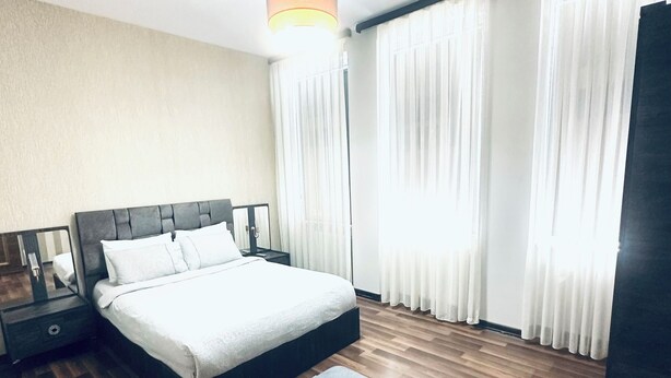 Quarto com cama, mesas de cabeceira, abajur, iluminação no teto, janela com cortinas e piso de madeira.