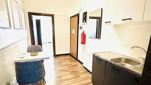 Um corredor estreito com piso de madeira, uma área de jantar com uma cadeira azul, uma cozinha com uma pia e um extintor de incêndio na parede.