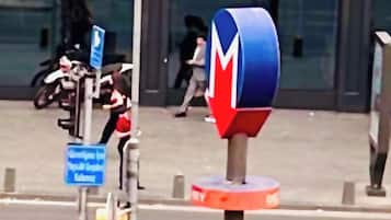 Una esquina con un letrero azul y rojo, un pequeño quiosco y un paso de peatones.