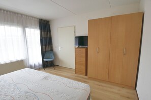 Ein Schlafzimmer mit Bett, Kleiderschrank, Fernseher auf einem Schrank, einem Sessel und einem Fenster mit Vorhängen.